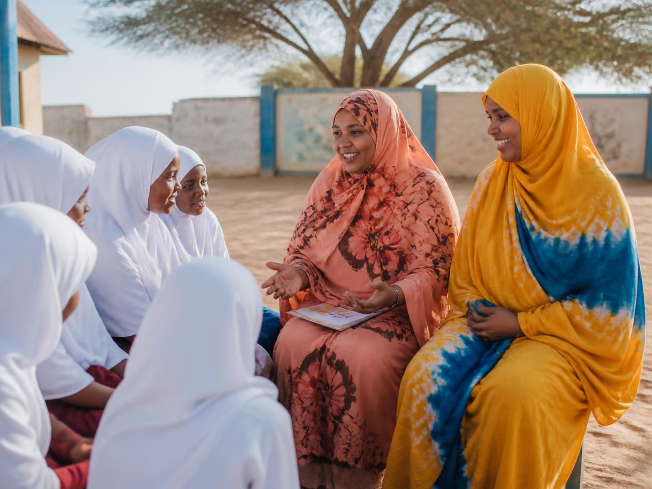 Somali women in their thirties and forties speaking with a group of schoolgirls in a sunlit courtyard